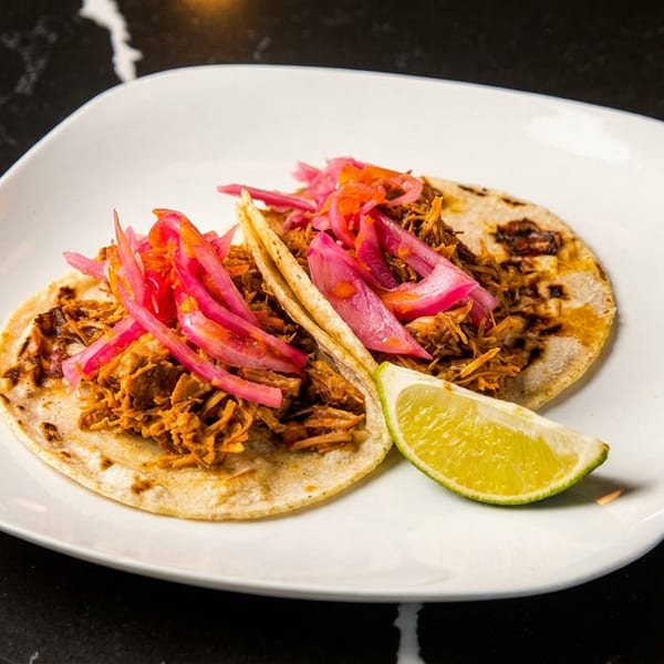 Two corn tortillas topped with shredded meat and pickled red onions, served with a lime wedge on a white plate.