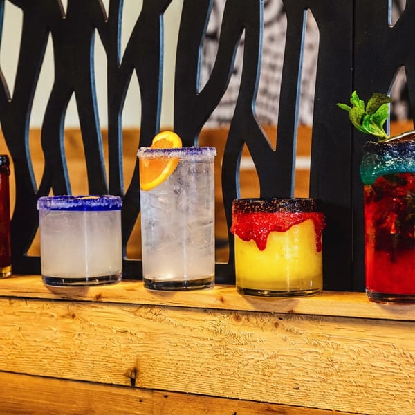 Five colorful cocktails with fruit garnishes and sugar-rimmed glasses displayed on a rustic wooden bar.