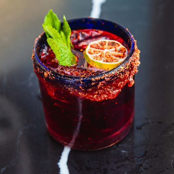 Deep red cocktail in a chili-salt-rimmed glass, garnished with mint leaves and a citrus wheel on a dark tabletop.