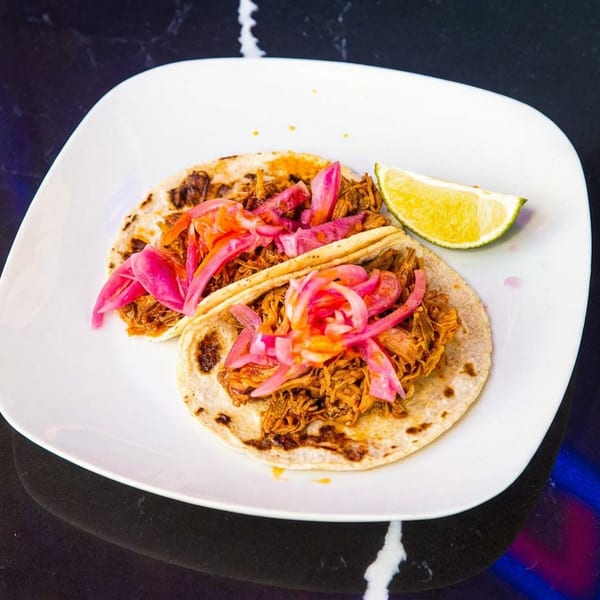 Two corn tortillas filled with shredded meat, pickled onions, and vegetables on a white plate with a lime wedge.