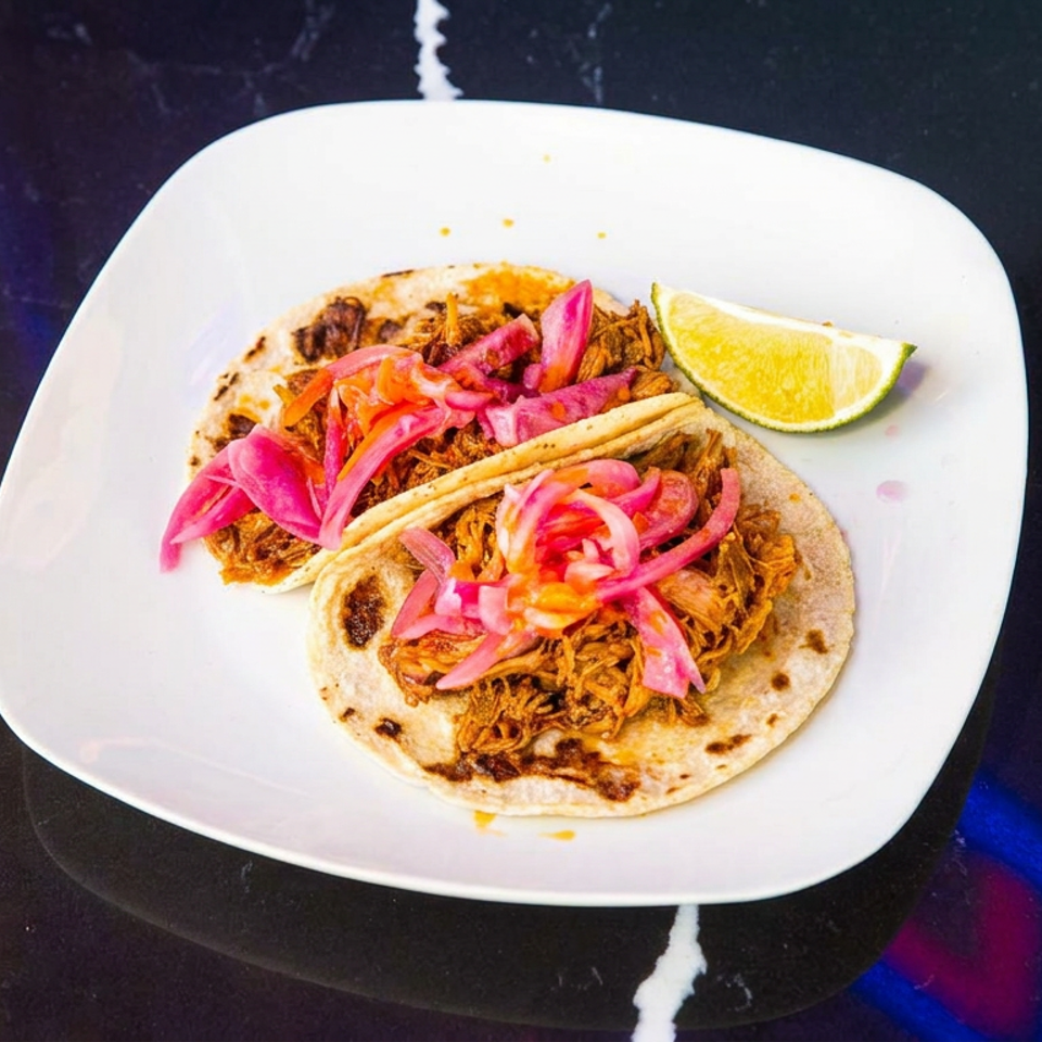 Two corn tortillas filled with shredded meat, pickled onions, and vegetables on a white plate with a lime wedge.