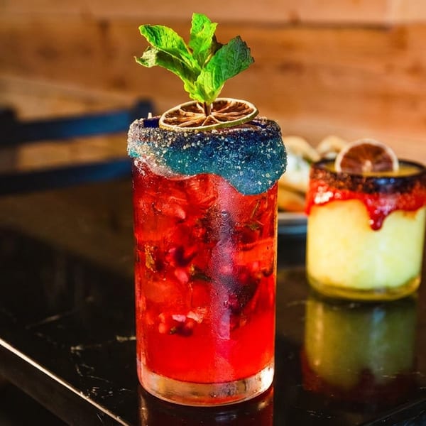 Colorful red and green cocktails with sugared rims, citrus slices, and mint garnishes on a reflective bar.