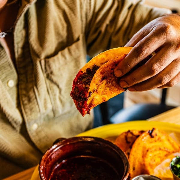 Diner holds a crispy folded taco filled with saucy meat over a bowl containing more tacos and dipping sauce.