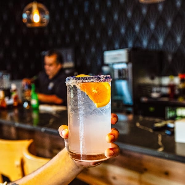 Hand holding a salted citrus cocktail with an orange slice in a warmly lit restaurant bar.