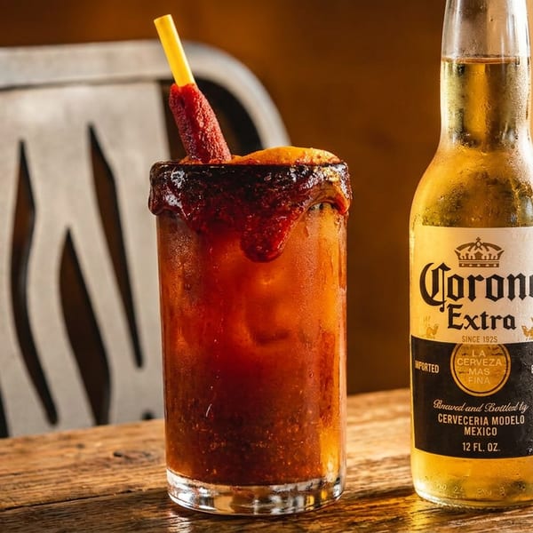 Spicy red beer cocktail garnished with a chili pepper beside a bottle of Corona Extra on a rustic wooden bar.