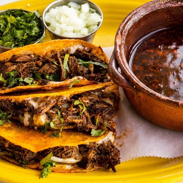 Three crispy birria tacos filled with shredded beef and cilantro, served with consommé, lime, onions, and herbs on a yellow plate.